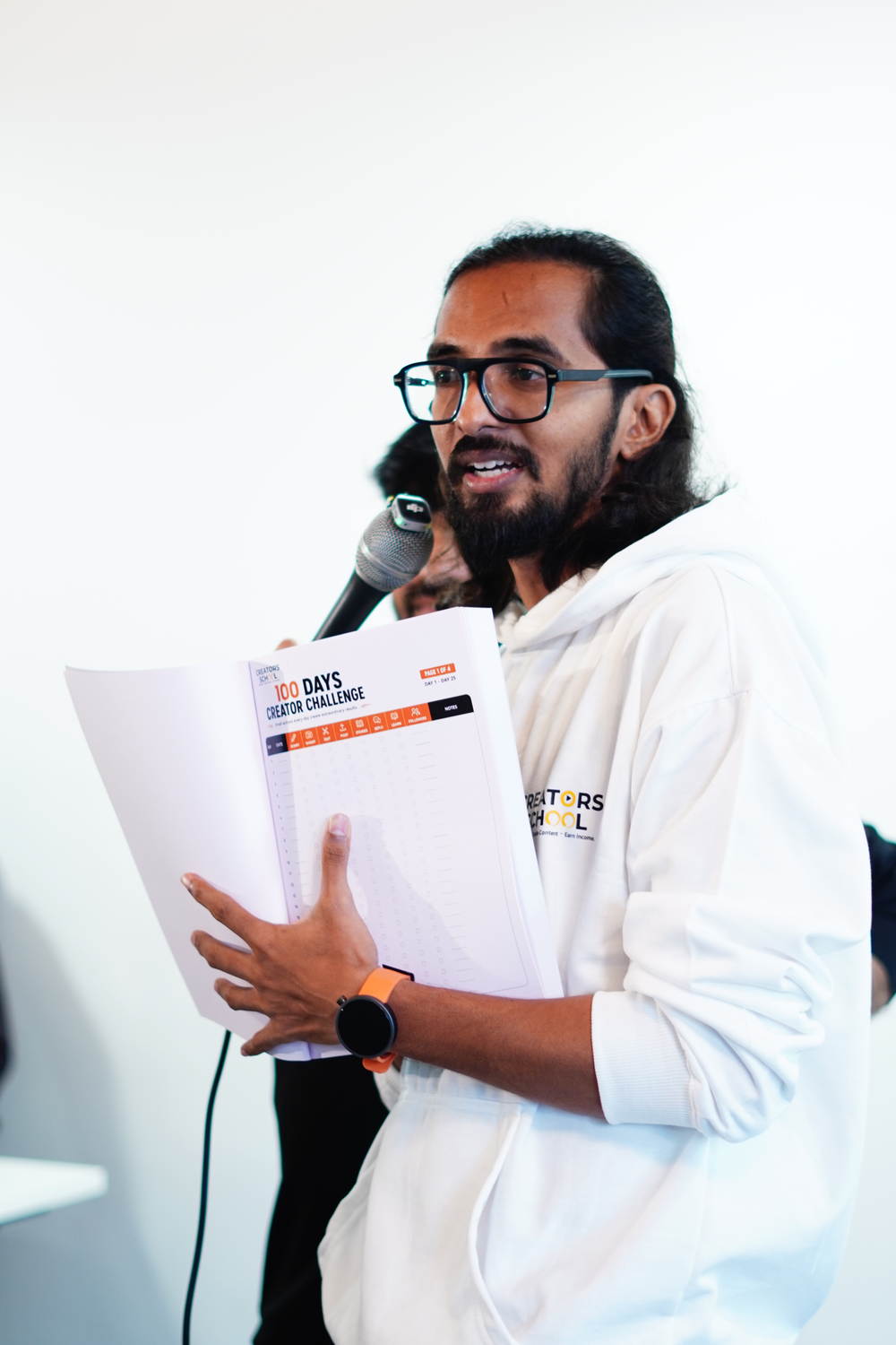 A student in a Creator's School hoodie holding the 15 day challenge sheet, speaking into a microphone